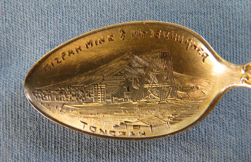 Souvenir Mining Spoon Mizpah Mine Bowl.JPG - SOUVENIR MINING SPOON MIZPAH MINE TONOPAH NV - Sterling silver souvenir demitasse spoon, features handle with miners and marked NEVADA, bowl withdetailed engraving of Mizpah mine scene and marked MIZPAH MINE & MT. BROUGHER at top and TONOPAH across bottom, length 4 1/8 in., marked on reverse Sterling and maker’s mark of S in a circle for Shepard Mfg. Co. MelroseHighlands, MA 1893-1923 (Mt. Brougher is a 6,500 ft. summit just to the west of Tonopah town) [Tonopah is an unincorporated town and the county seat of Nye County, Nevada. It is located approximately midway between Las Vegas and Reno.  One of the richest booms in the west occurred at Tonopah Springs on May 19, 1900. And the name Jim Butler will forever be associated with the name Tonopah and the many stories that surround the discovery. The legendary tale of discovery says that he went looking for a burro that had wandered off during the night and sought shelter near a rock outcropping. When Butler discovered the animal the next morning, he picked up a rock to throw at it in frustration, noticing that the rock was unusually heavy. He had stumbled upon the second-richest silver strike in Nevada history. News of the discovery traveled to the Klondike and soon scores of eager prospectors were searching the area. But it was not until August 27, 1900 that Butler and his wife filed on eight claims near the springs, six of which were some of the biggest producers the state has ever had including the Desert Queen, Burro, Valley View, Silver Top, Buckboard, and Mizpah, the largest silver producer in the district. Because the Butler claims were known far and wide, the town was often referred to as Butler. By the summer of 1901, the mines around the town produced nearly $750,000 worth of gold and silver. Now it was time for a post office and one opened on April 10, 1901 named Butler.  By 1902 Jim Butler had sold his claims, which were all consolidated and gave birth to a new company, the Tonopah Mining Company. It was incorporated in Delaware, with stock listed on both the Philadelphia and San Francisco exchanges. The company, with J.H. Whiteman as president, controlled 160 acres of mineral-bearing ground around the Tonopah district. The company also had holdings in the Tonopah-Goldfield Railroad and controlled mining companies in Colorado, Canada, California and Nicaragua. The mine workings at Tonopah consisted of three deep shafts with more than 46 miles of lateral workings. The deepest of the three shafts was 1,500 ft. The ore mined at the site was treated in a 100-stamp mill. Also in 1902 the Tonopah-Belmont Mining Company was formed and was based in New Jersey with C.A. Heller as president. The company’s property, 11 claims covering more than 160 acres, was on the east side of the property owned by the Tonopah Mining Company. There were two deep vertical shafts, 1,200 and 1,700 ft, with workings covering almost 39 miles.  Butler now had a population of 650 and was increasing every day. It also had six saloons, restaurants, assay offices, lodging houses, and a number of doctors and lawyers. It was not until March 3, 1905 that its name was changed to Tonopah. By 1907, Tonopah had become a full-fledged city with modern hotels, electric and water companies, five banks, schools, and hundreds of other buildings. Tonopah’s mines continued to produce extremely well until the Depression brought a slowdown. From 1900 to 1921, they produced ore worth almost $121 million. Tonopah’s biggest year was 1913 when its mines yielded almost $10 million worth of gold, silver, copper, and lead. By the time World War II started, only four major mining companies were operating in Tonopah. At the end of the war even the companies that had been there at the beginning were gone. In 1968, Howard Hughes and his Summa Corporation bought 100 claims in Tonopah, including the Mizpah, Silver Top, and Desert Queen mines. Hopes for a mining revival soon faded after disappointing core samples were taken. A few of the old mines were re-timbered but never reopened. The value of the Tonopah district’s total production is just over $150 million.  Today tourism plays a large part in the local economy.]